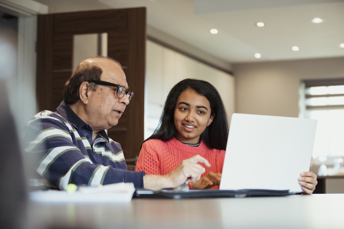 A grandfather is helping his granddaughter as she works from a laptop; they look happy to be bonding and working together.