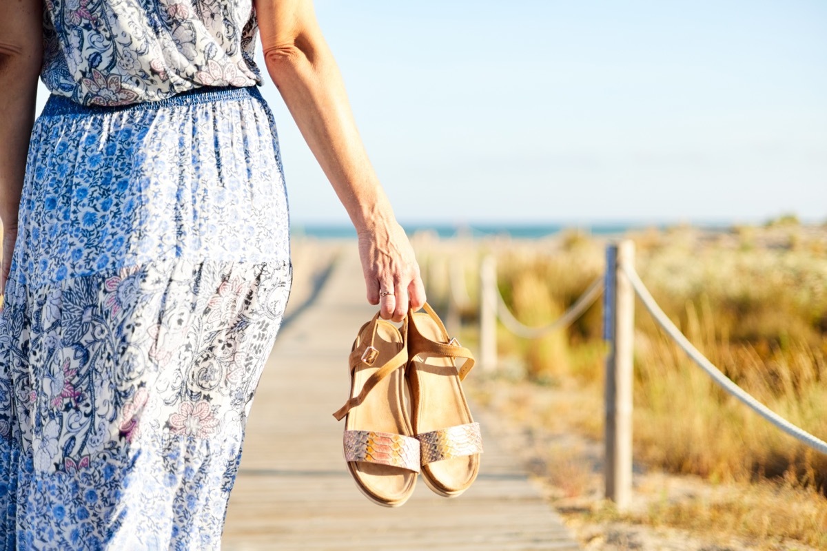 Middle aged woman walking on the beach with sandals in her hand. She is wearing a flowery summer dress.