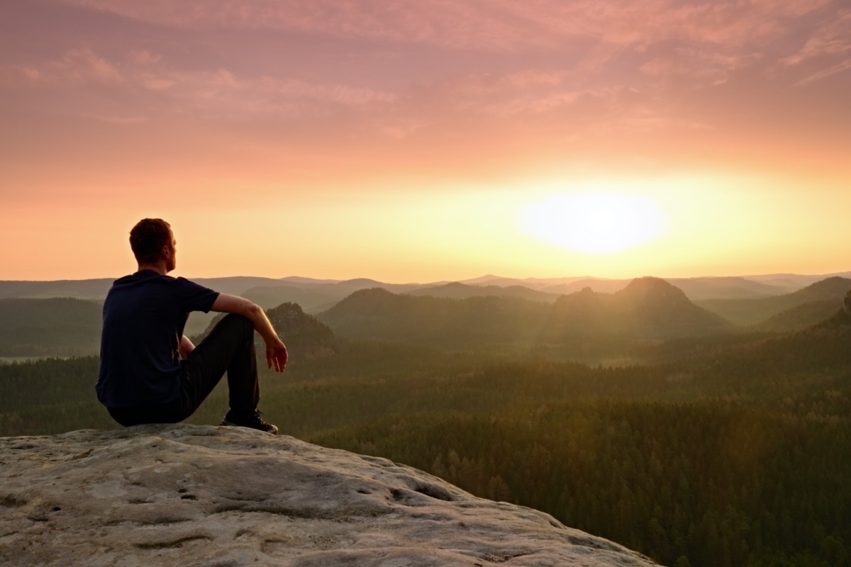 Tourist in squatting position on peak of sandstone rock and watching into colorful mist and fog in morning valley