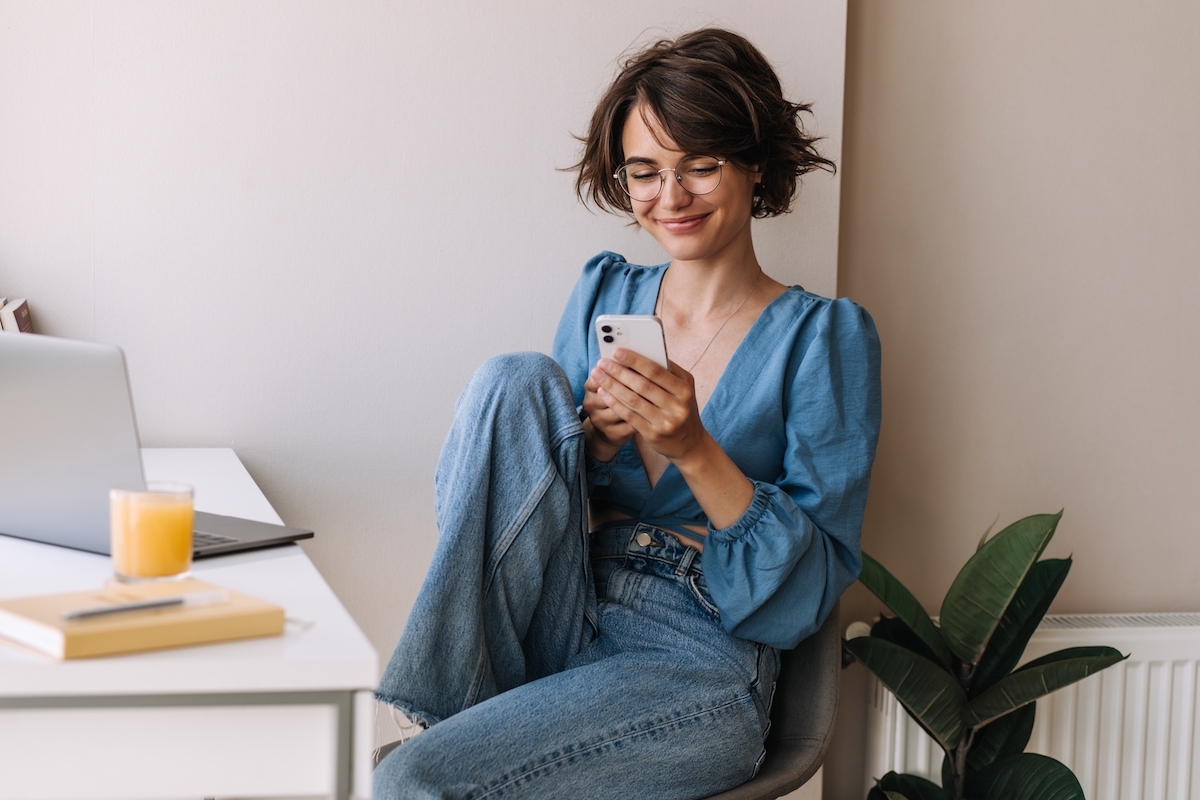 Young woman with a bob hairstyle wearing a blue blouse and jeans sits at her desk using her phone