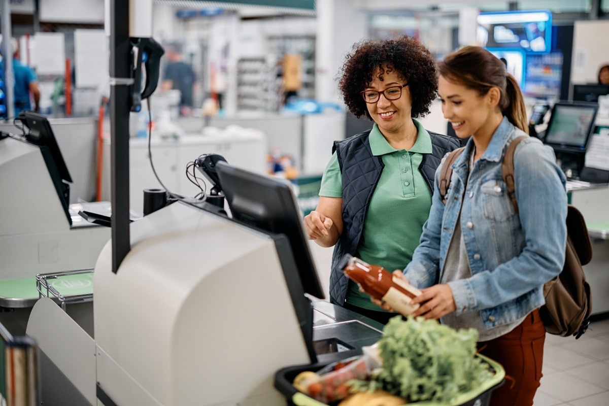 Young happy woman using self-service checkout with help of supermarket worker.