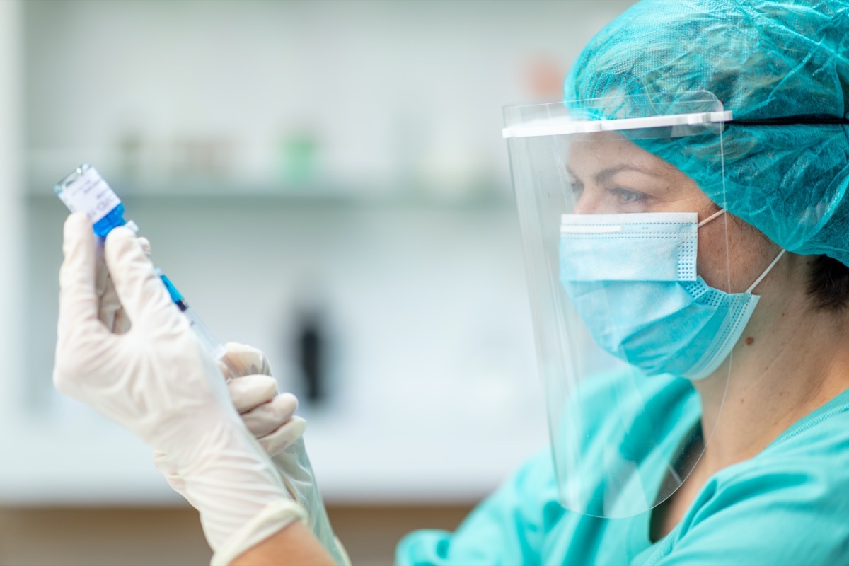 Female doctor holding COVID-19 vaccine vial and taking liquid solution out with syringe; prevention and immunization from corona virus infection.