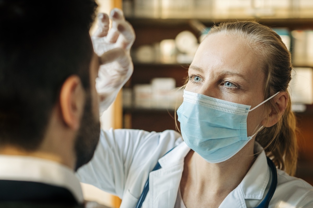 Female healthcare worker wearing surgical mask examining male patient. Doctor checking eyes of mid adult man. They are at a hospital.