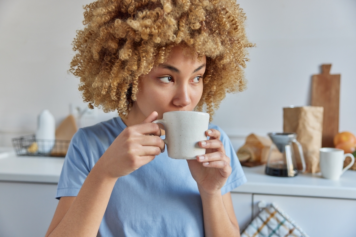 A young woman with curly hair wearing a sky-blue t-shirt drinks coffee in her kitchen