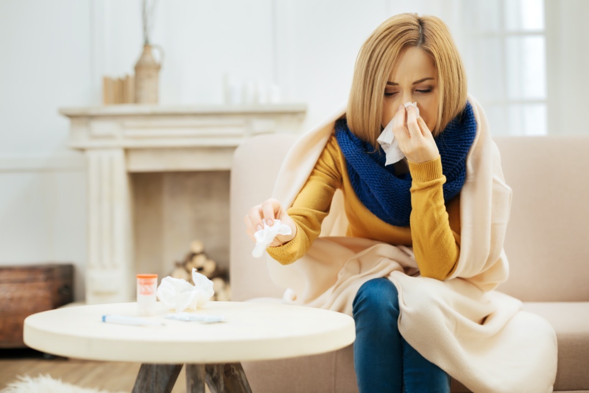 Ill young blond woman having fever and blowing her nose while having a blanket on her shoulders and sitting on the couch with her eyes closed and table with pills in front of her