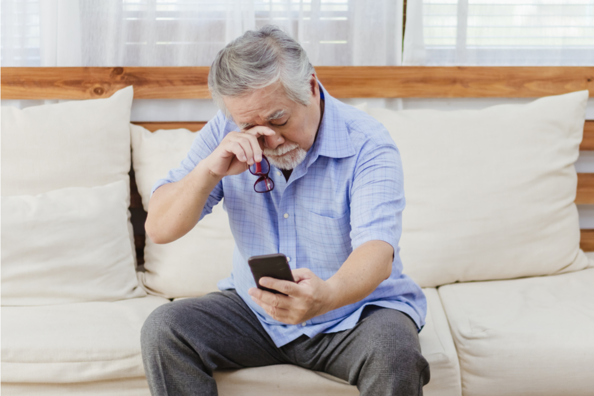 man sitting on couch holding phone outstretched and rubbing his eyes