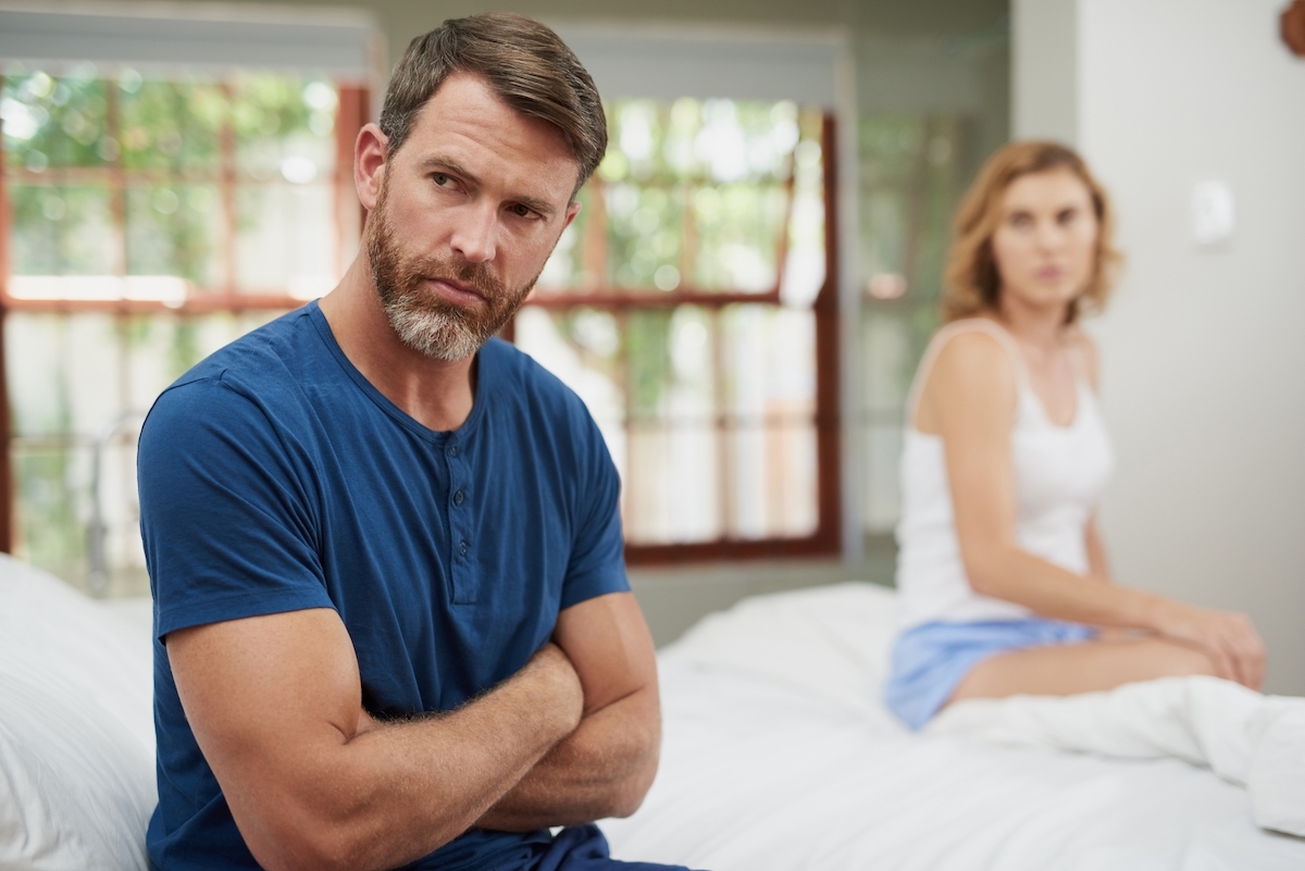 An upset man sitting on the bed with his arms folded in the foreground with his wife looking on in the background.