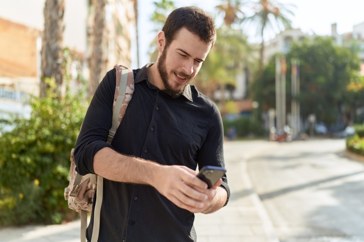 Young man with a beard wearing a black shirt and backpack texting while walking down the street