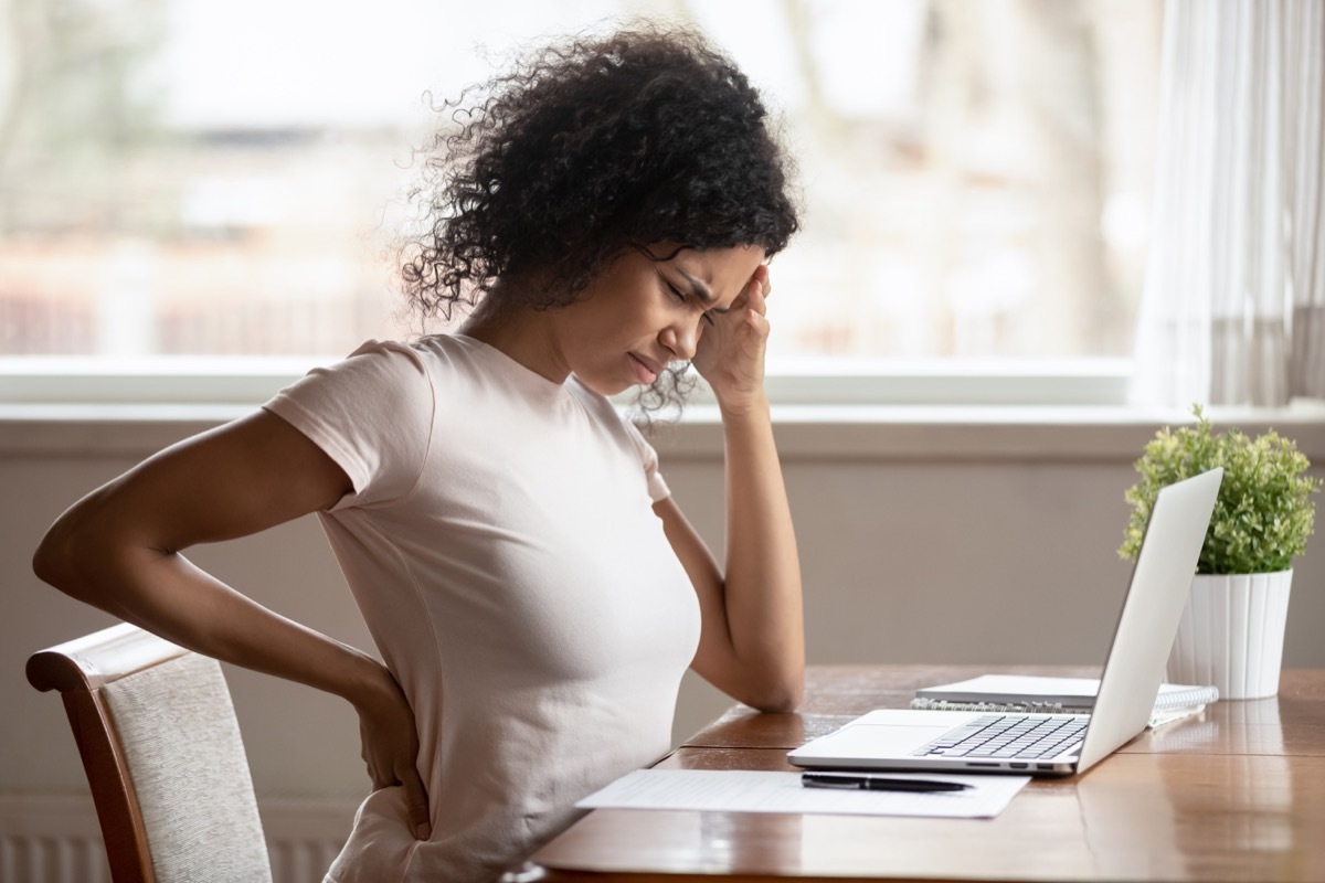 woman looking uncomfortable, hand on back and head, back pain near desk