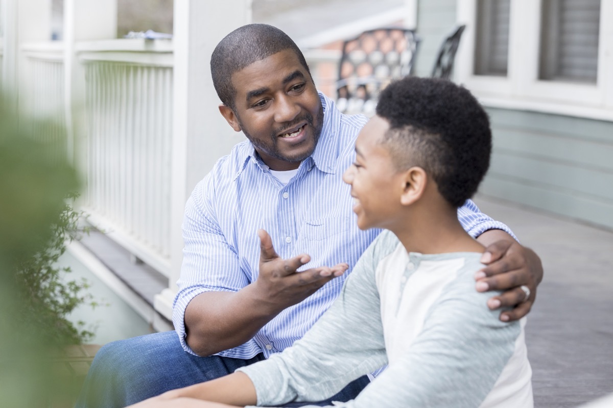 An excited mid adult father sits on the front steps of his home with his preteen son and gestures as they enjoy an exciting conversation.