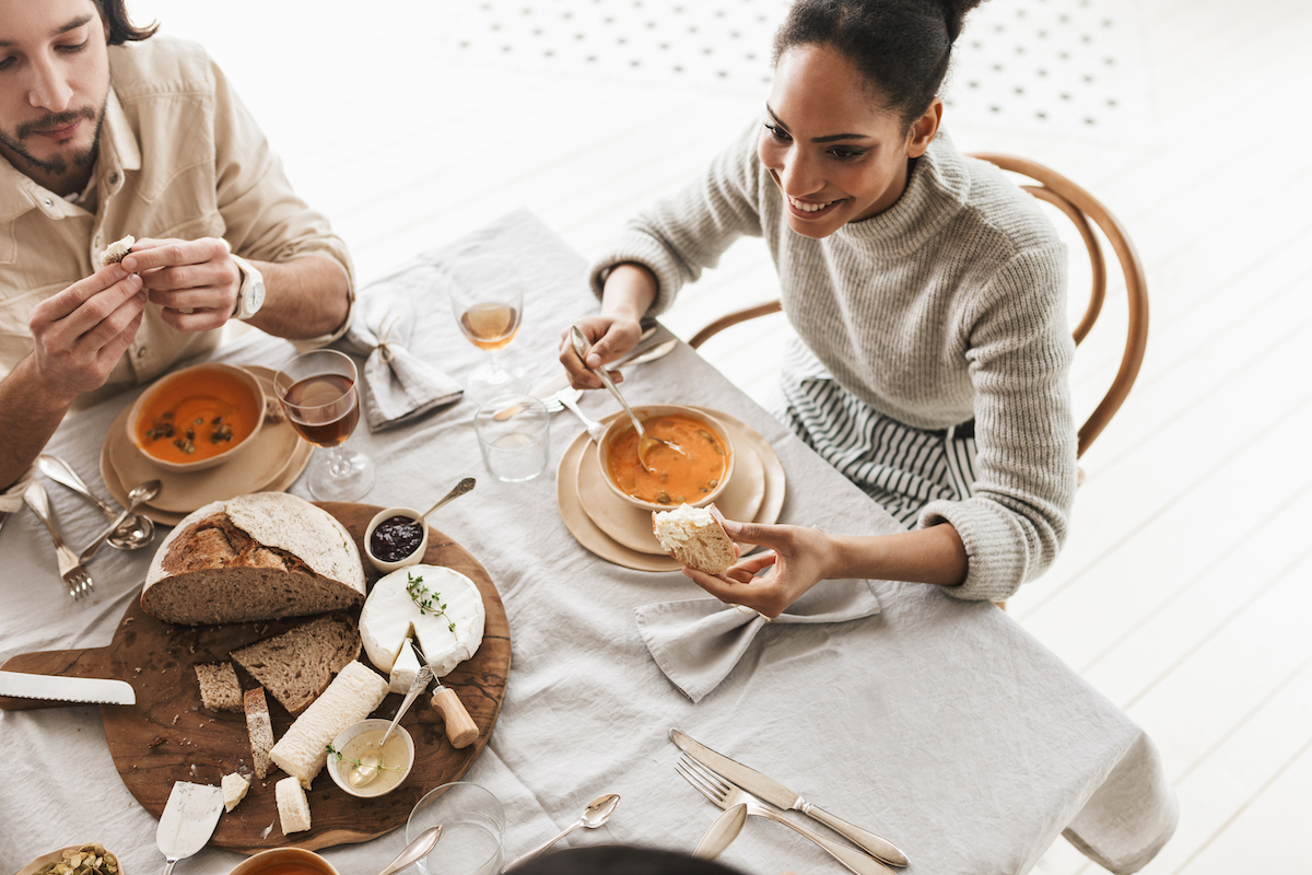 Top view of a couple dressed in neutrals eating soup, along with bread and cheese.