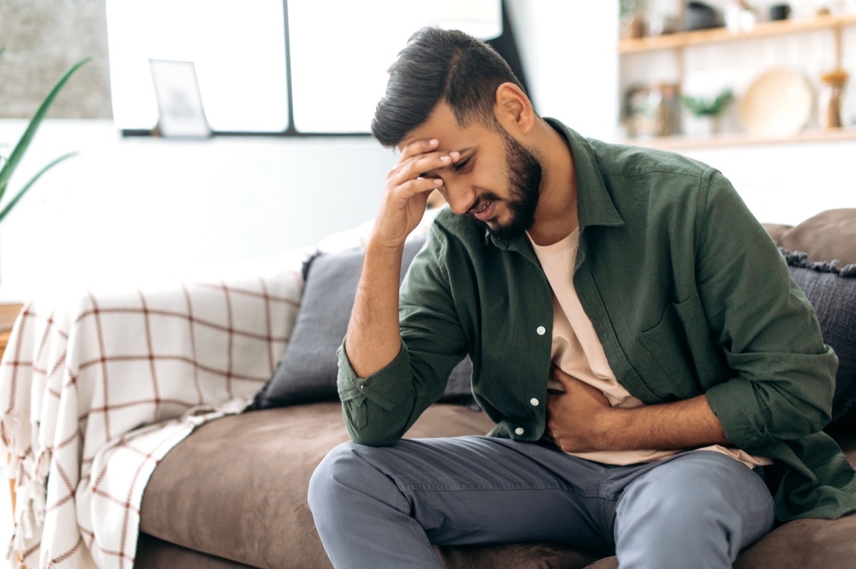 Unhappy indian man sits on comfortable sofa in cozy living room, holds his hands on his stomach, grimaces from pain in his stomach