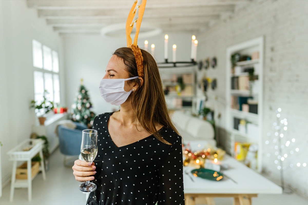 Photo of a young woman holding a glass of champagne, wearing a protective face mask and reindeer antlers; celebrating and hosting a Christmas dinner party at home during the COVID-19 pandemic.