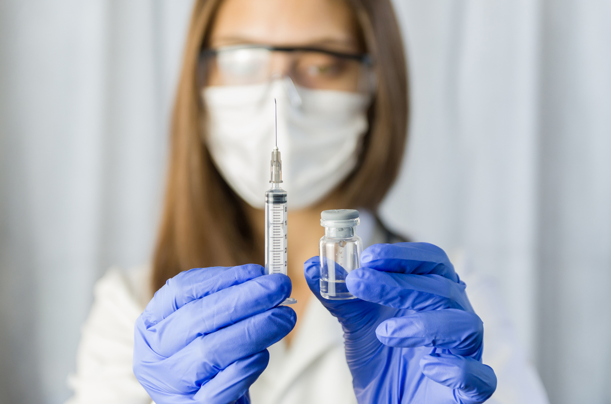 A female healthcare worker wearing a face mask, protective goggles, and gloves holds a syringe and vial of COVID-19 vaccine