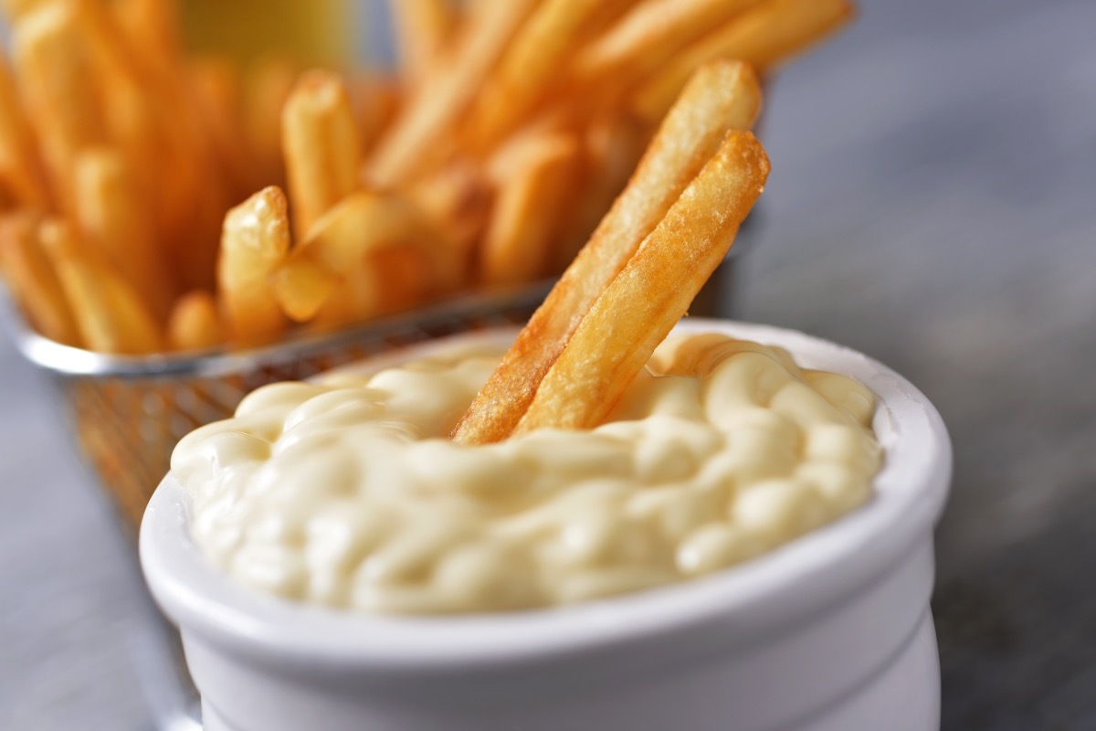 two french fries dipped into a white ramekin of mayonnaise with a container of fries in the background