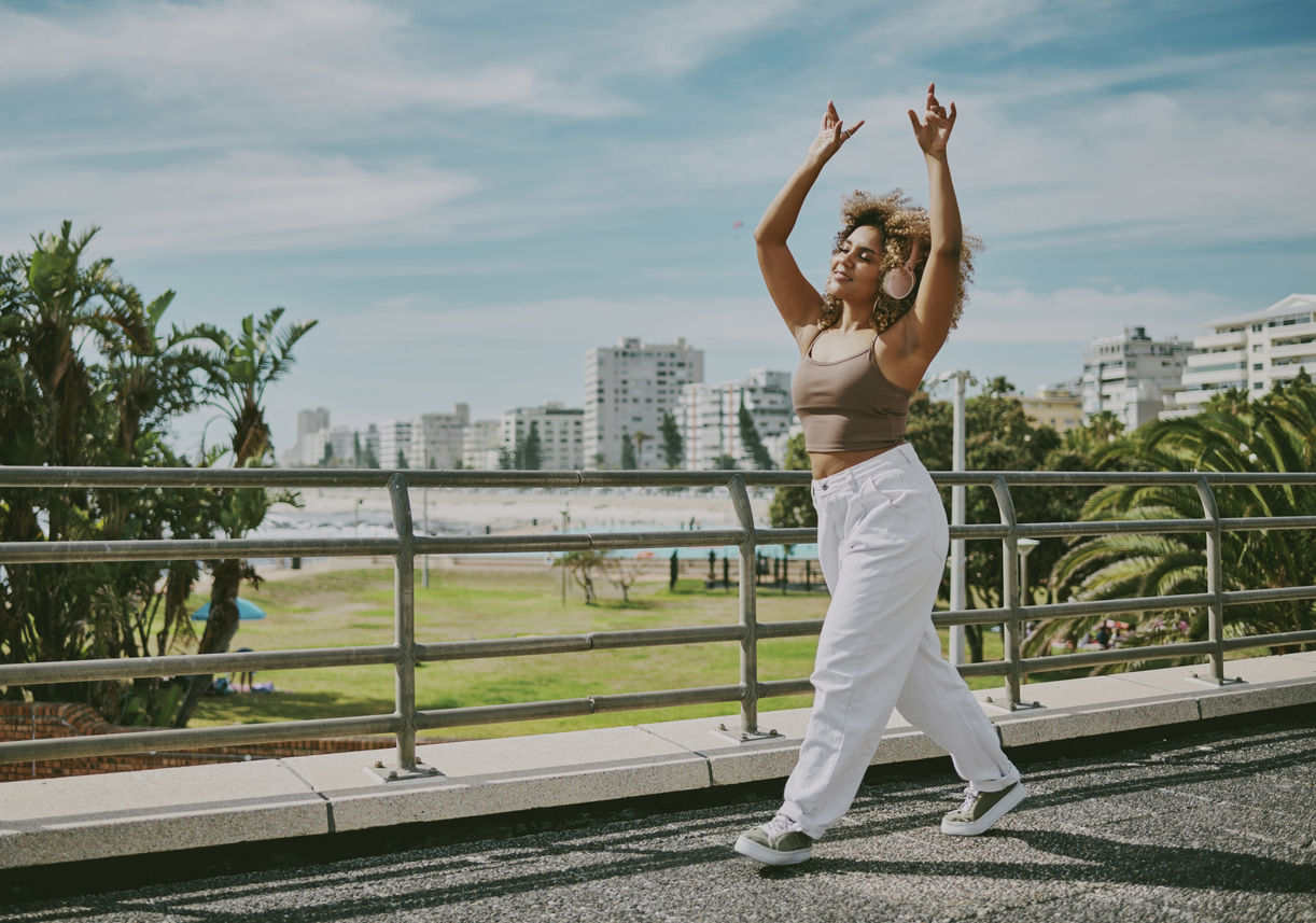 young woman listening to music with her hands in the air while taking a walk outside near the beach