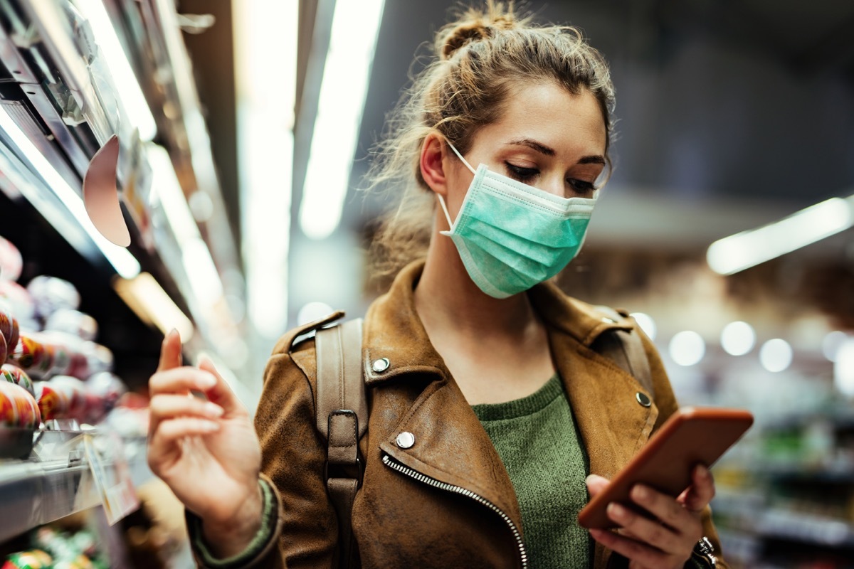 Young woman wearing protective mask on her face and reading shopping list on mobile phone in grocery store during virus pandemic.