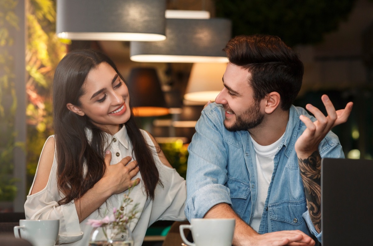 Man and woman flirting at a cafe; the woman is touching the man's back and holding her other hand to her chest