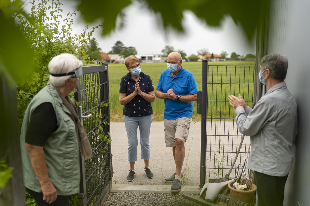 two senior couples , friends meeting after many weeks of covid-19 lockdown, curfew, quarantine, all wearing mouth nose masks or protective face shield, greeting with social distance at garden fence door in rural village (two senior couples , friends m