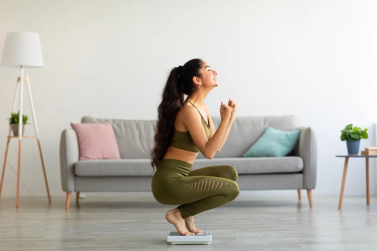 Full length of excited Indian woman sitting on scales at home, overjoyed with success of her slimming diet, side view. Emotional Asian lady achieving her weight loss goal, making YES gesture