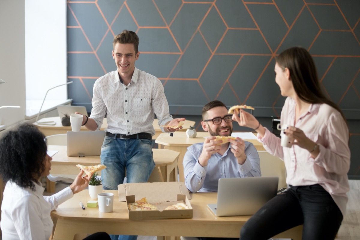 Friendly diverse team talking and laughing eating pizza together, happy colleagues sharing meal, multiracial coworkers group enjoying lunch at break, good relations and office food delivery