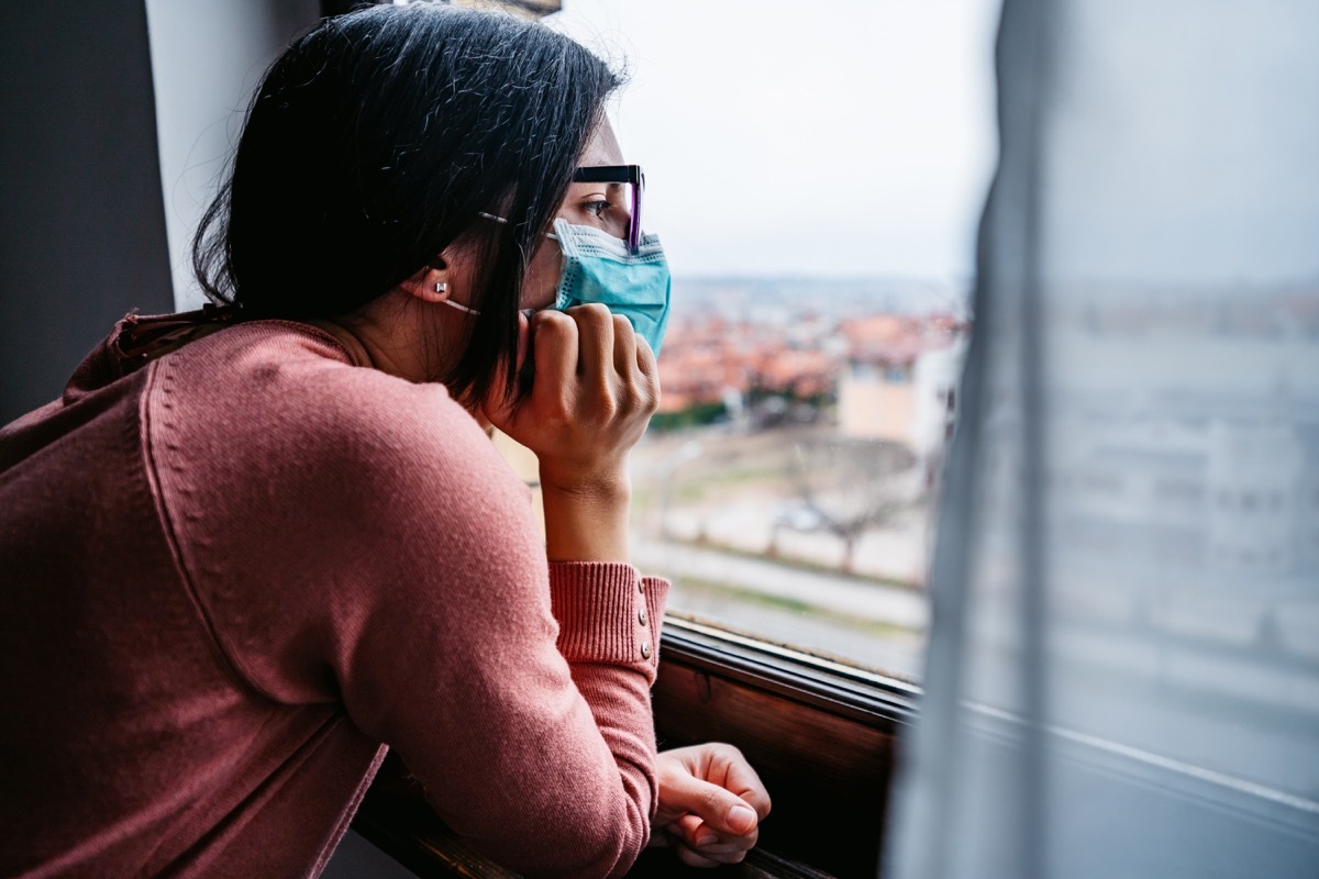 Woman in quarantine wearing a mask and looking through the window. Pandemic of Coronavirus / Covid-19.