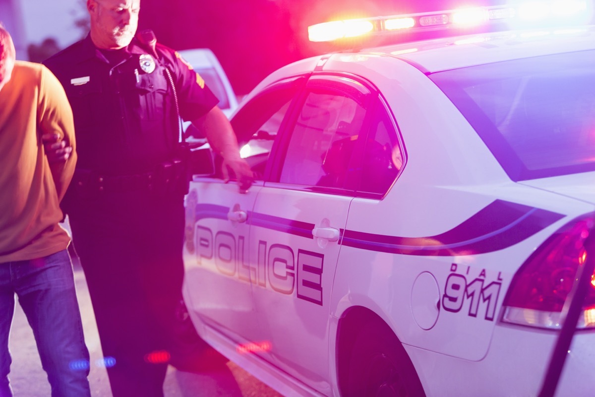 Police officer making an arrest, escorting a young man in handcuffs toward the back seat of his police car, at night.