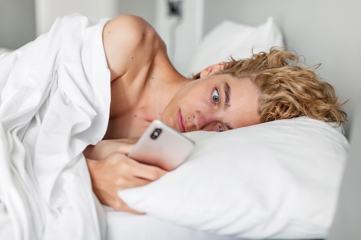 Surprised and concerned man waiting for a phone call while lying on bed and looking at cell phone. Sad teenager read the bad news