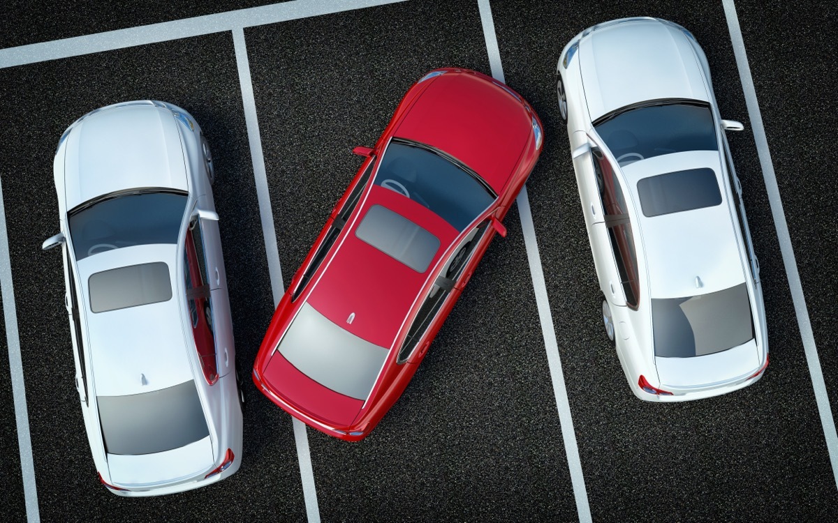 Red car parked over the lines in a parking lot with two white cars next to it