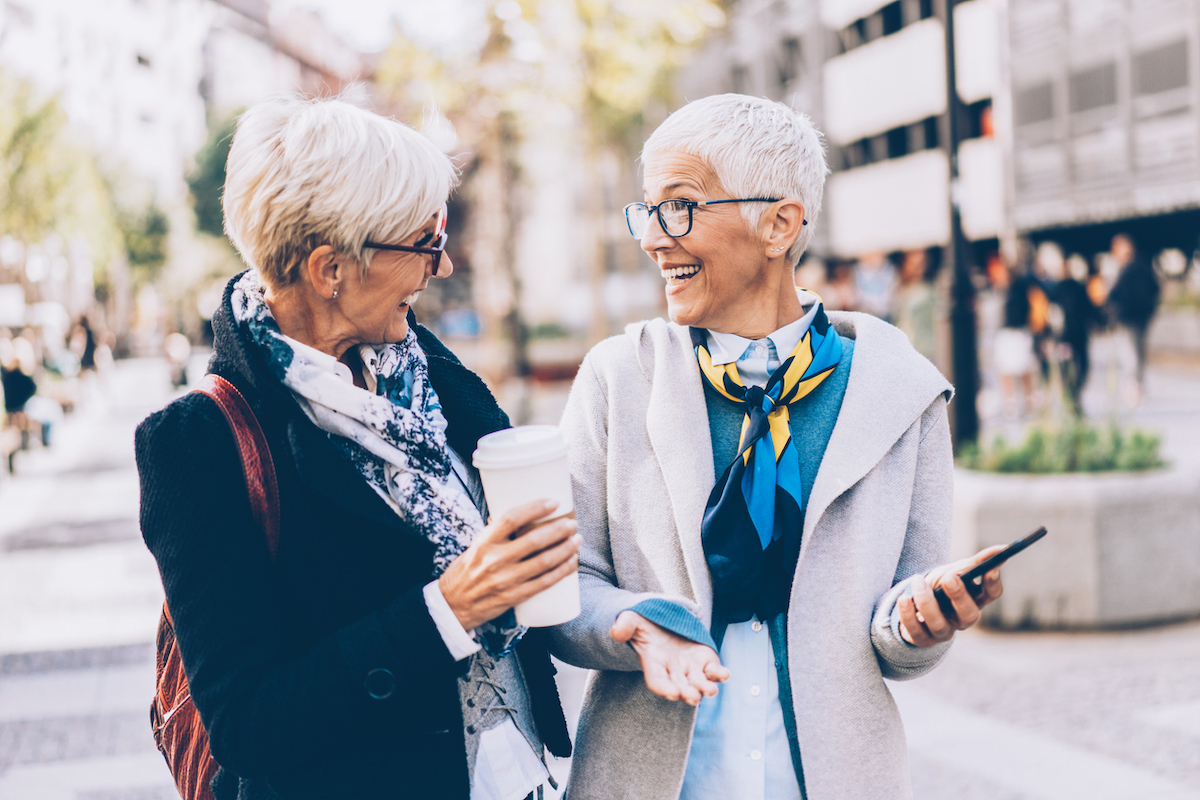 Two senior women, both with short white hair and wearing jackets and scarves, walk down the street laughing.