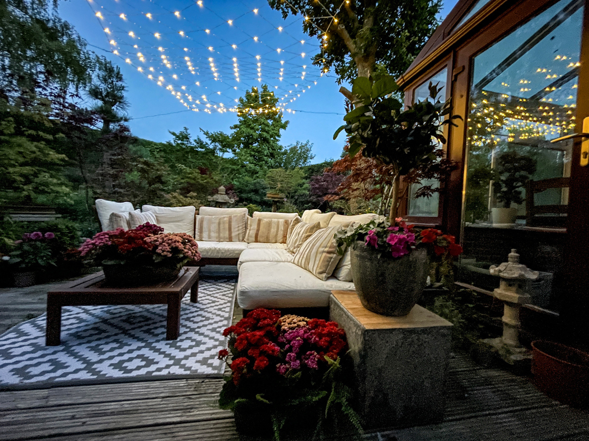 Stock photo showing ornamental Japanese-style garden with outdoor lounge area at night illuminated with fairy lights. Featuring crystal clear koi pond, whitewashed, grooved timber decking patio, Japanese bonsai maples and hardwood, cushion covered seating.