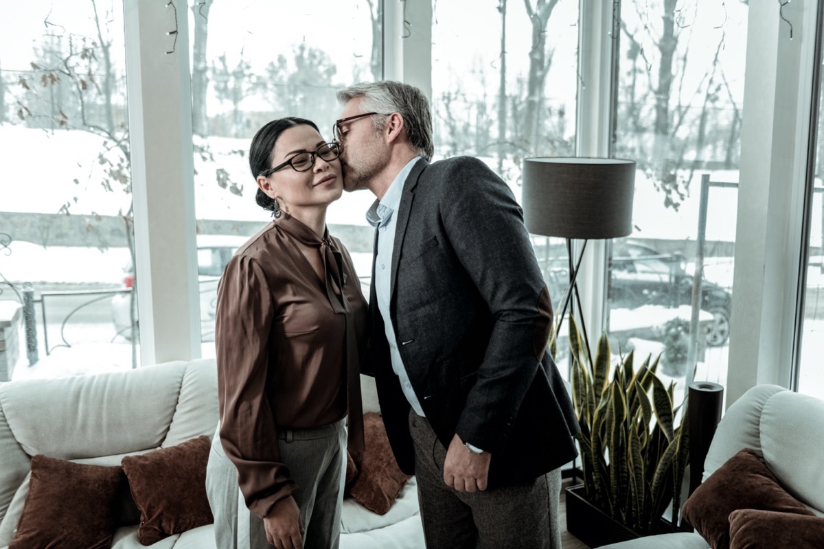 Kiss on a cheek. Tall grey-haired handsome man wearing a blue shirt kissing a woman on a cheek