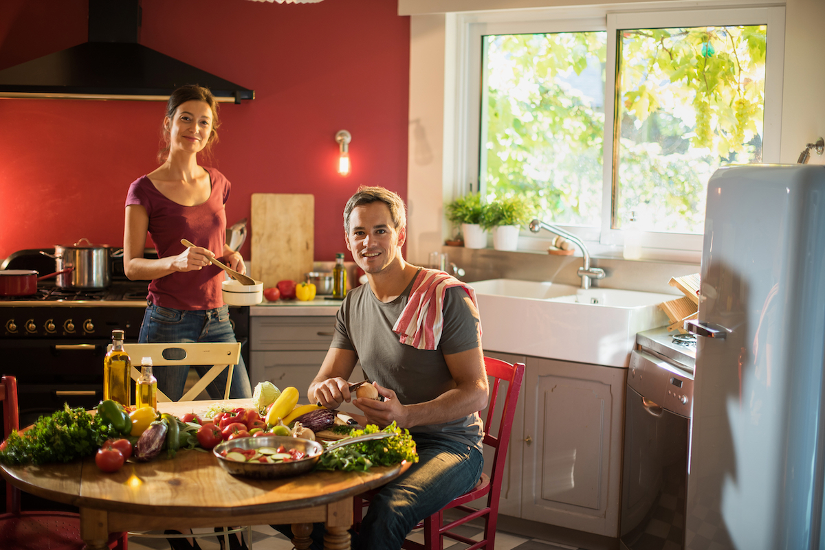 Couple smiling in a kitchen while making dinner. The kitchen has a red wall and a gray, retro fridge.