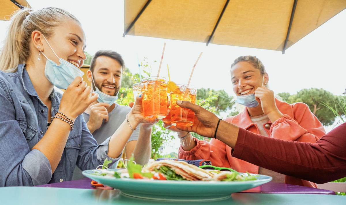 A group of friends drinking together at an outdoor bar while sharing food and lowering their face masks