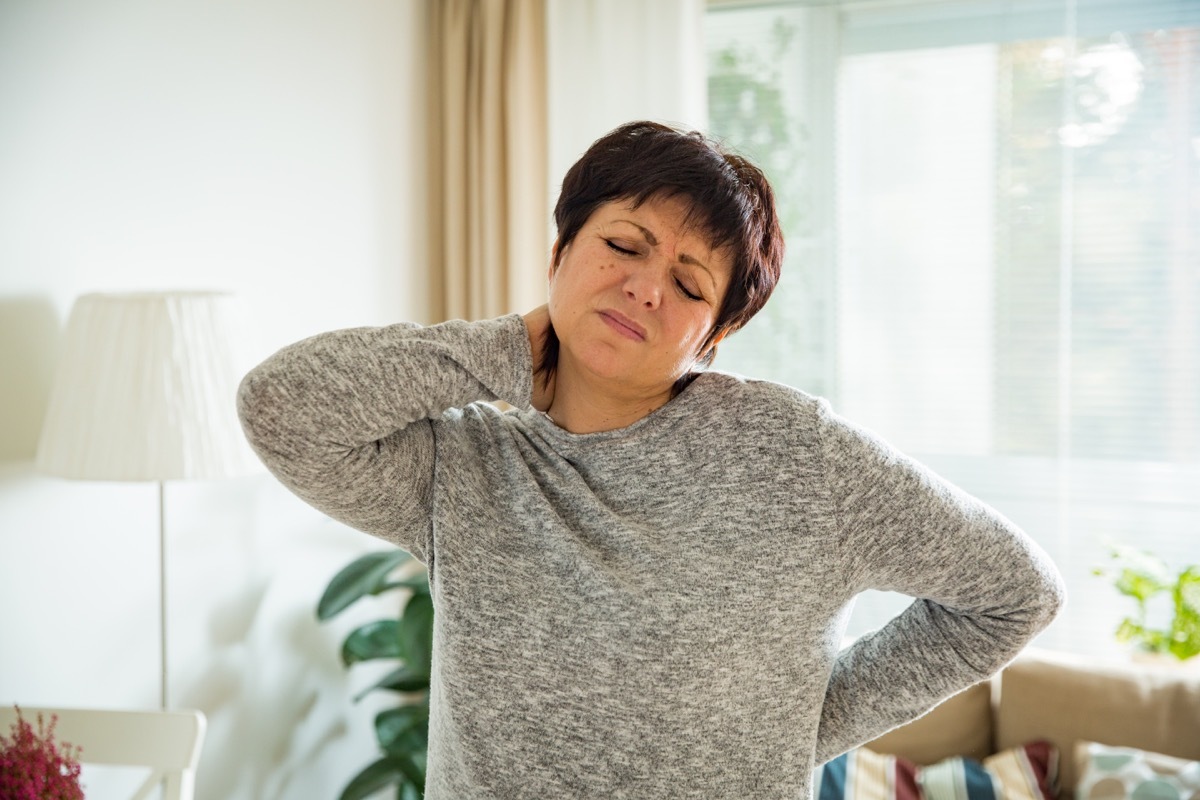 Mature woman suffering from backache at home. Massaging neck with hand, feeling exhausted, standing in living room.