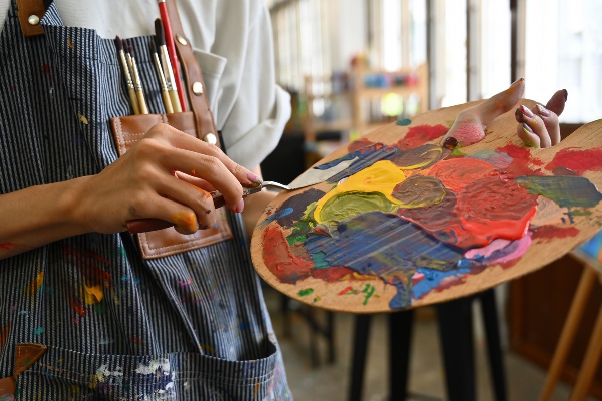 Close-up image of a young creative artist's hands smeared with watercolors holding a spatula mixing color on a palette.