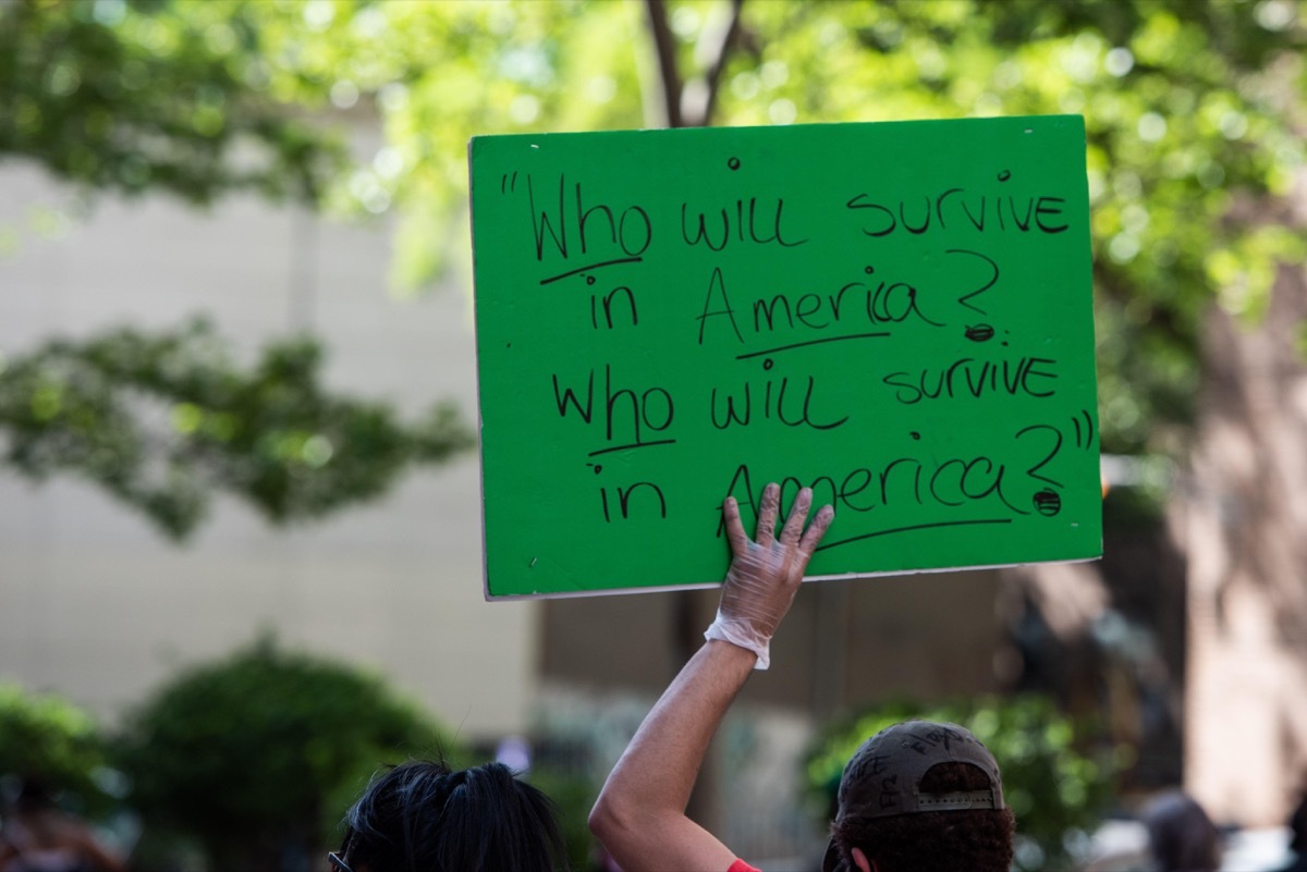 2BWD77M Protestor marching against the police killing of George Floyd carrying a sign saying Who will survive in America? Who will survive in America? quoting a spoken word poem by Gil Scott-Heron.