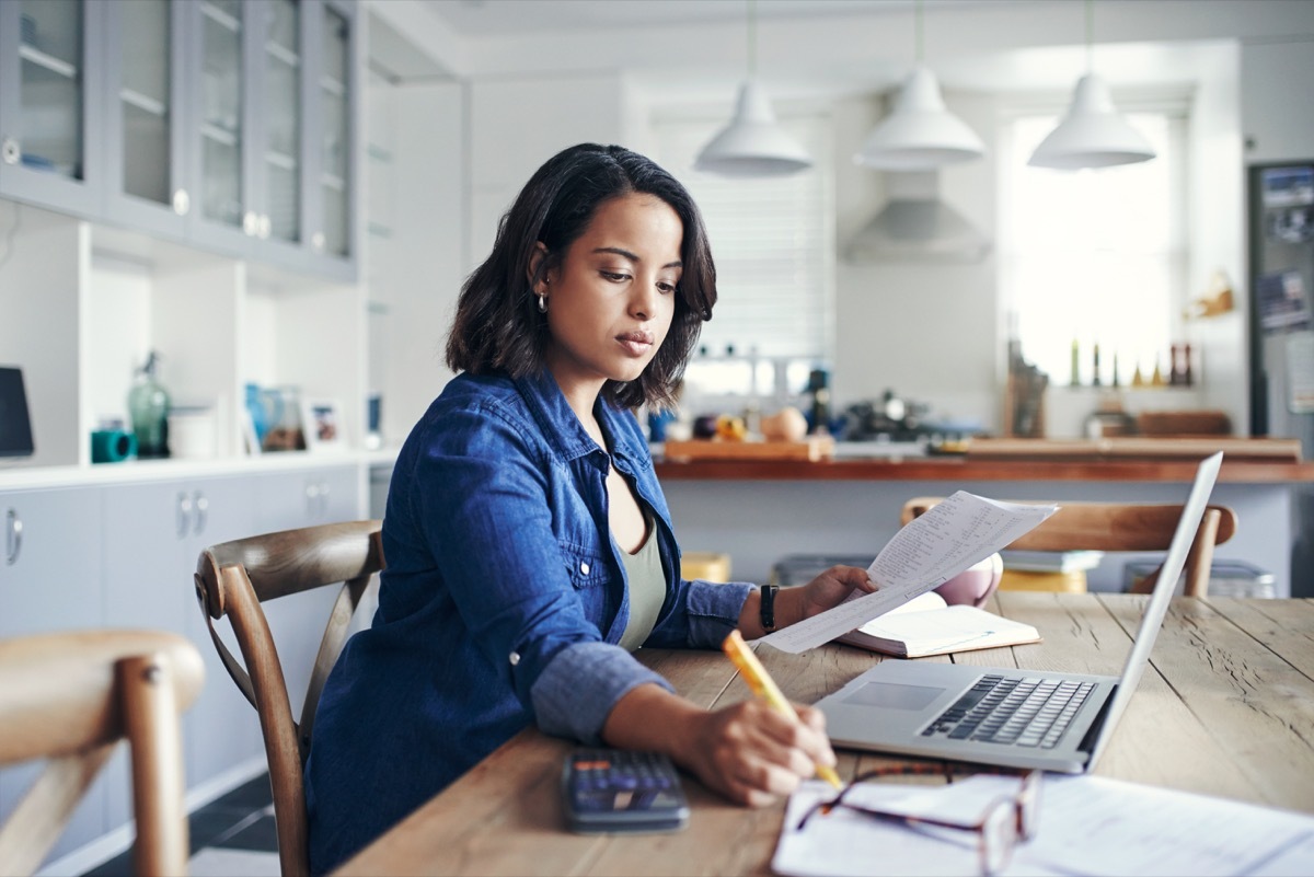 Shot of a young woman using a laptop and going through paperwork while working from home