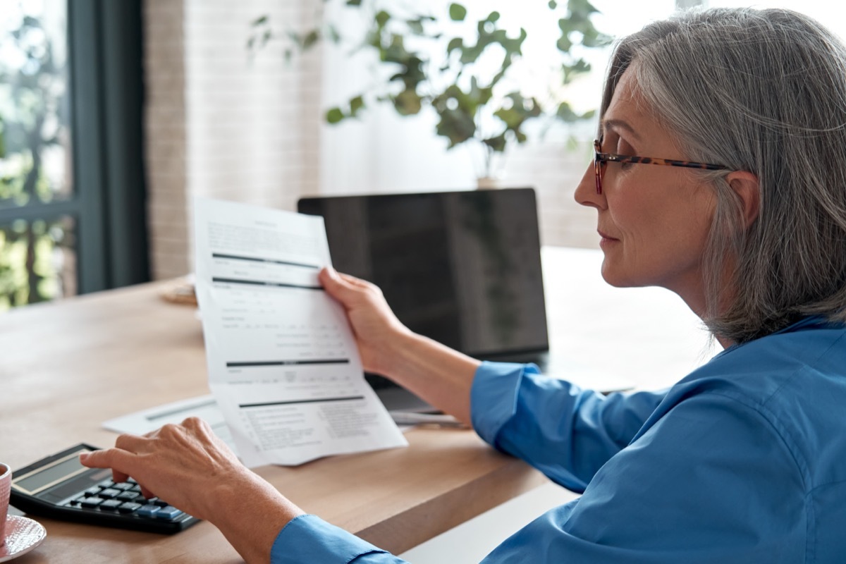 mature woman holding a paper bill using a calculator at her desk