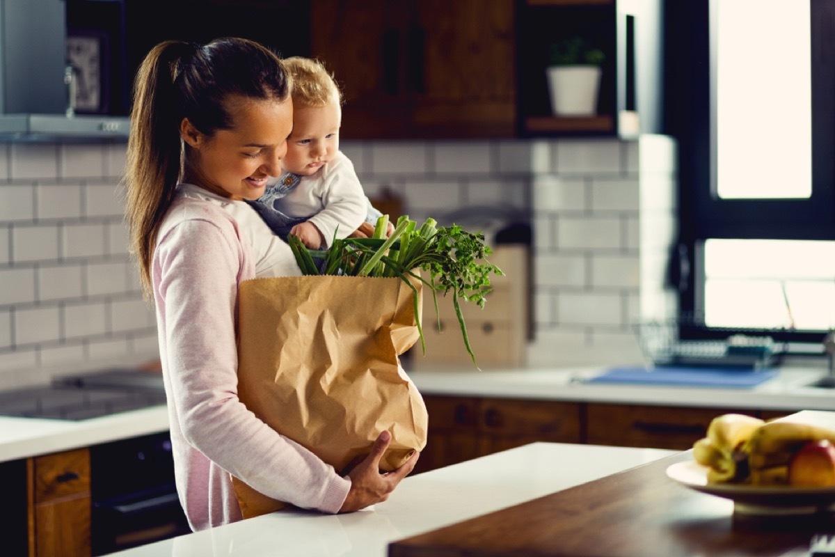 white woman holding baby and a bag of groceries in the kitchen