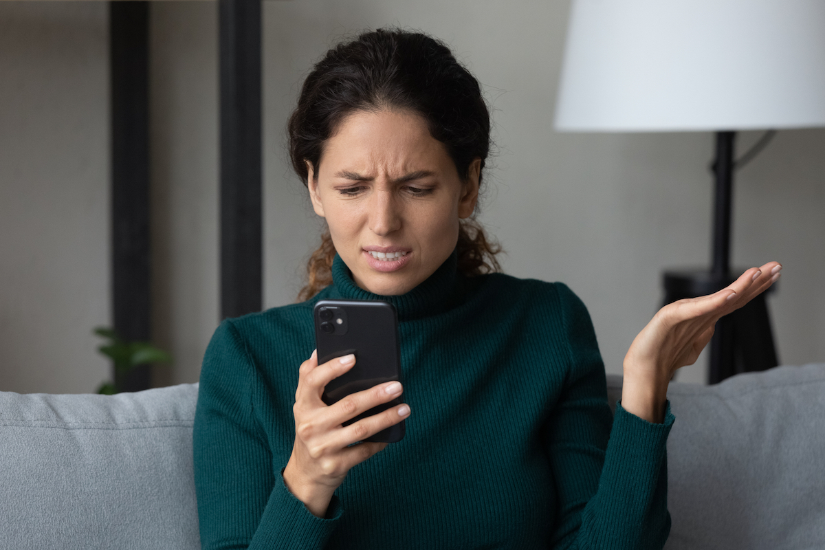 A young woman sits on her couch and looks at her phone annoyed.