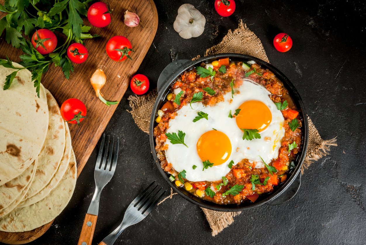 Traditional Mexican dish Huevos rancheros - scrambled eggs with tomato salsa, with taco tortillas, fresh vegetables and parsley. Breakfast for two. Top view.