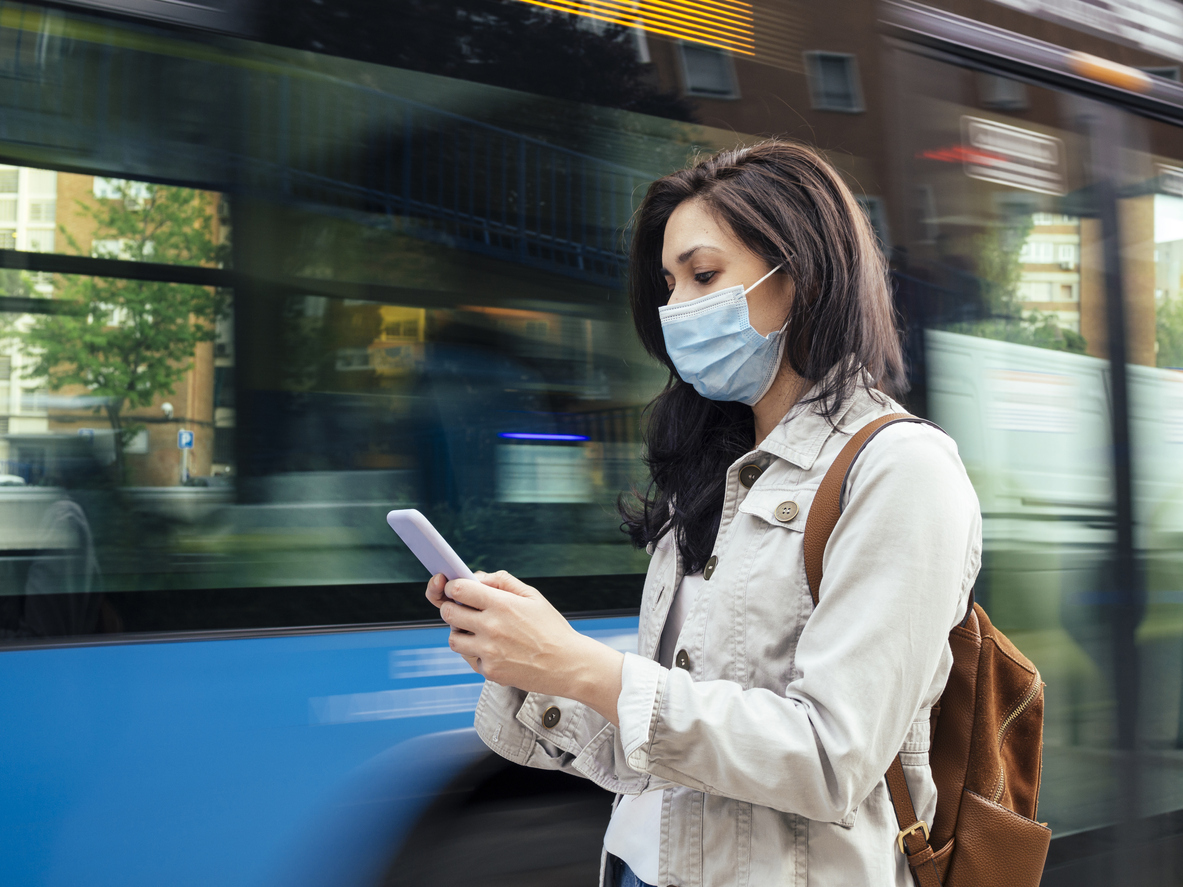 A young woman wearing a face mask checks her smartphone while waiting for a city bus