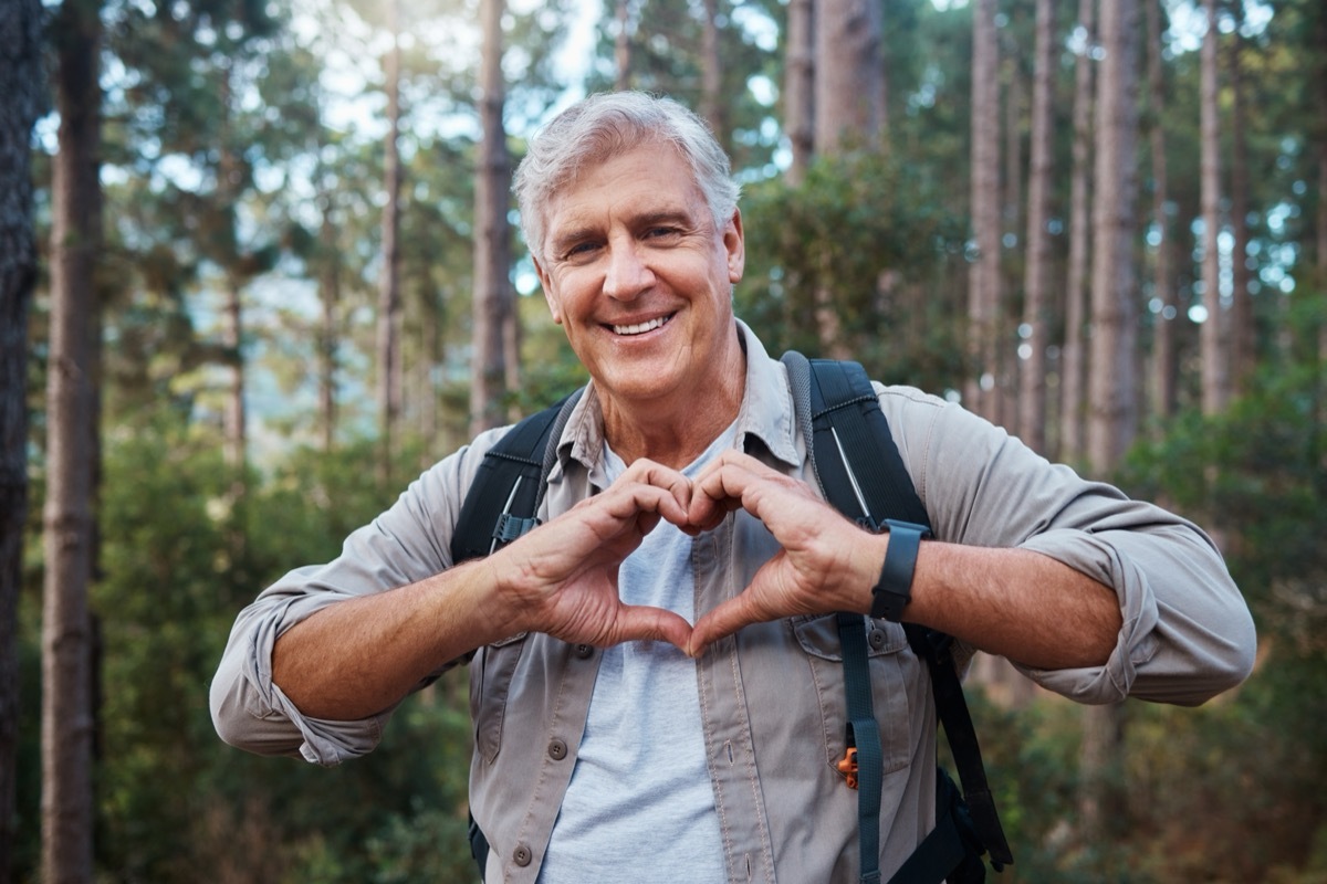 Portrait, heart and senior man in forest hiking, fitness and health support, self care hands and cardiovascular workout. Nature, travel or trekking of happy person in woods with love sign or emoji