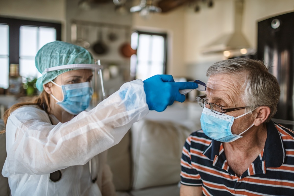 Female doctor using thermometer to measure the temperature of senior man for Covid-19 testing at home.