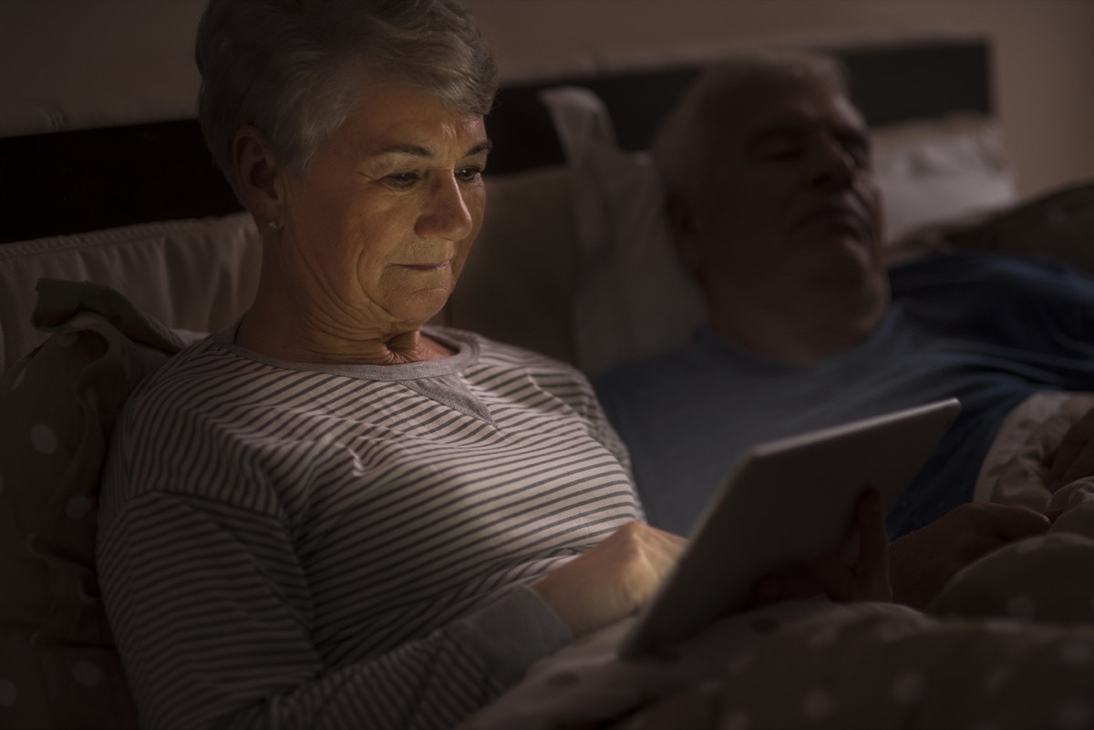 older white woman using ipad in bed while husband sleeps next to her