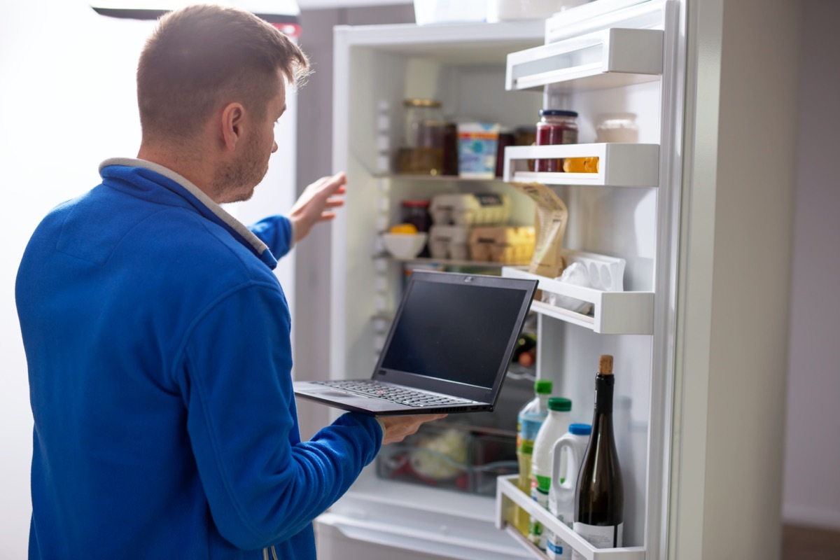 Man holding laptop and reaching into refrigerator