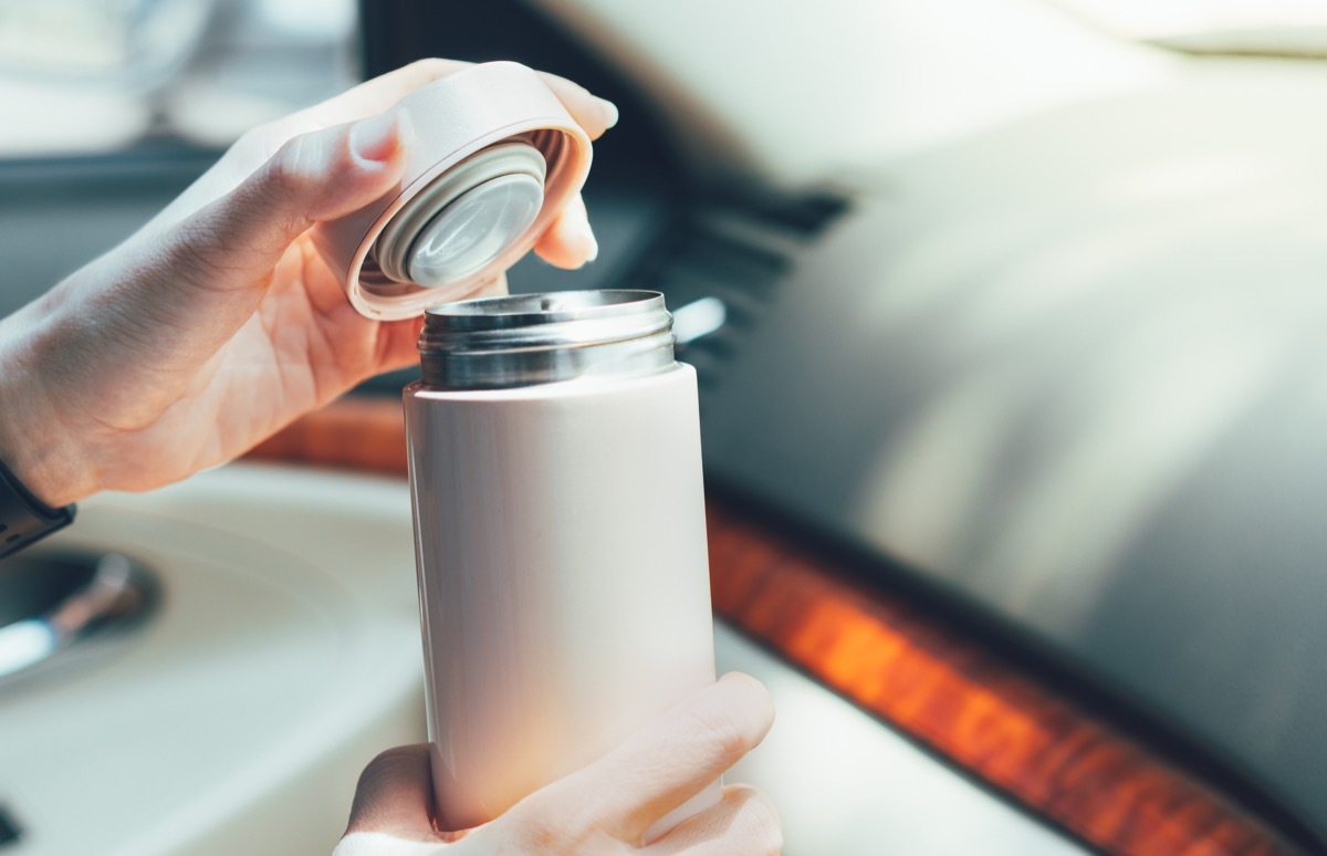 Closeup, Woman's hands hold insulated reusable water bottle in car instead of single use plastic cup, open lid to drink her coffee. Eco friendly, zero waste and green living lifestyle.