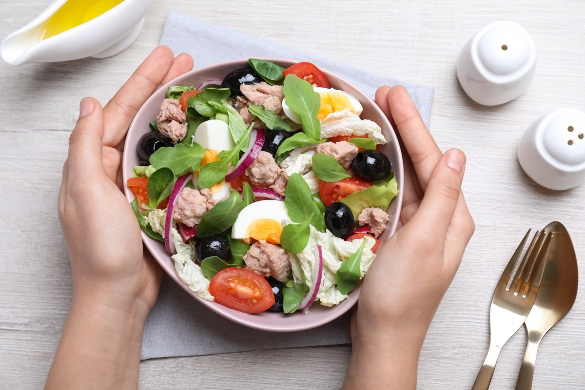 Woman holding bowl of delicious salad with canned tuna at white wooden table, top view